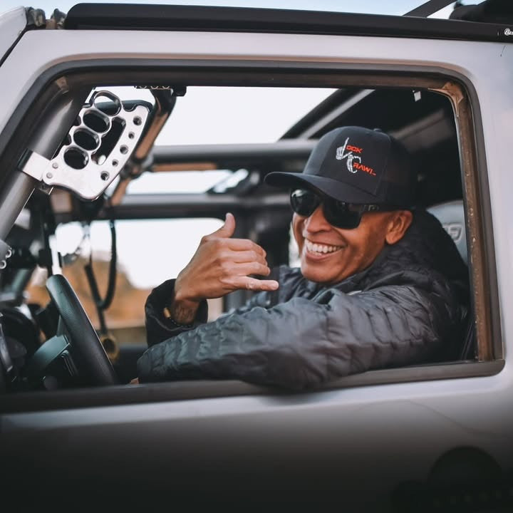 Person sitting inside a vehicle, wearing a cap and sunglasses, with a blurred background.